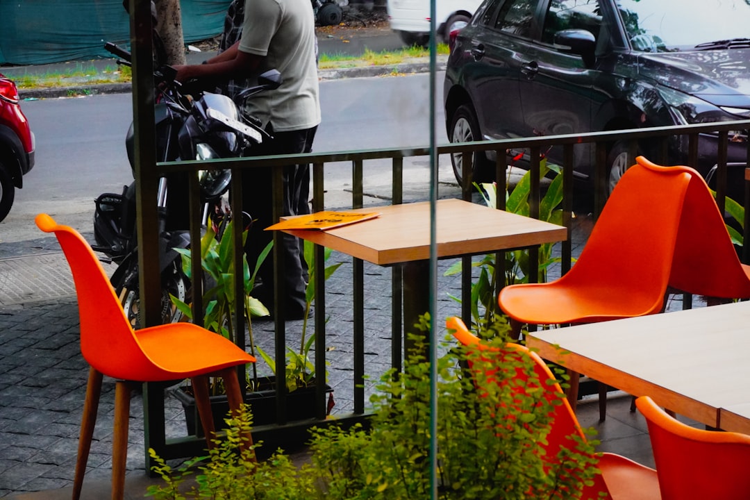 Outdoor cafe tables with bright orange chairs, the established side of the asymmetry, where months of evidence have already wired the entity into multiple surfaces